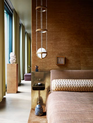 un lit en laiton avec une tête de lit en tissu et un couvre-lit assorti, le mur derrière recouvert d'un tissu or ambré, une table en marbre et cuir à côté du lit et une suspension en laiton, verre et daim au-dessus